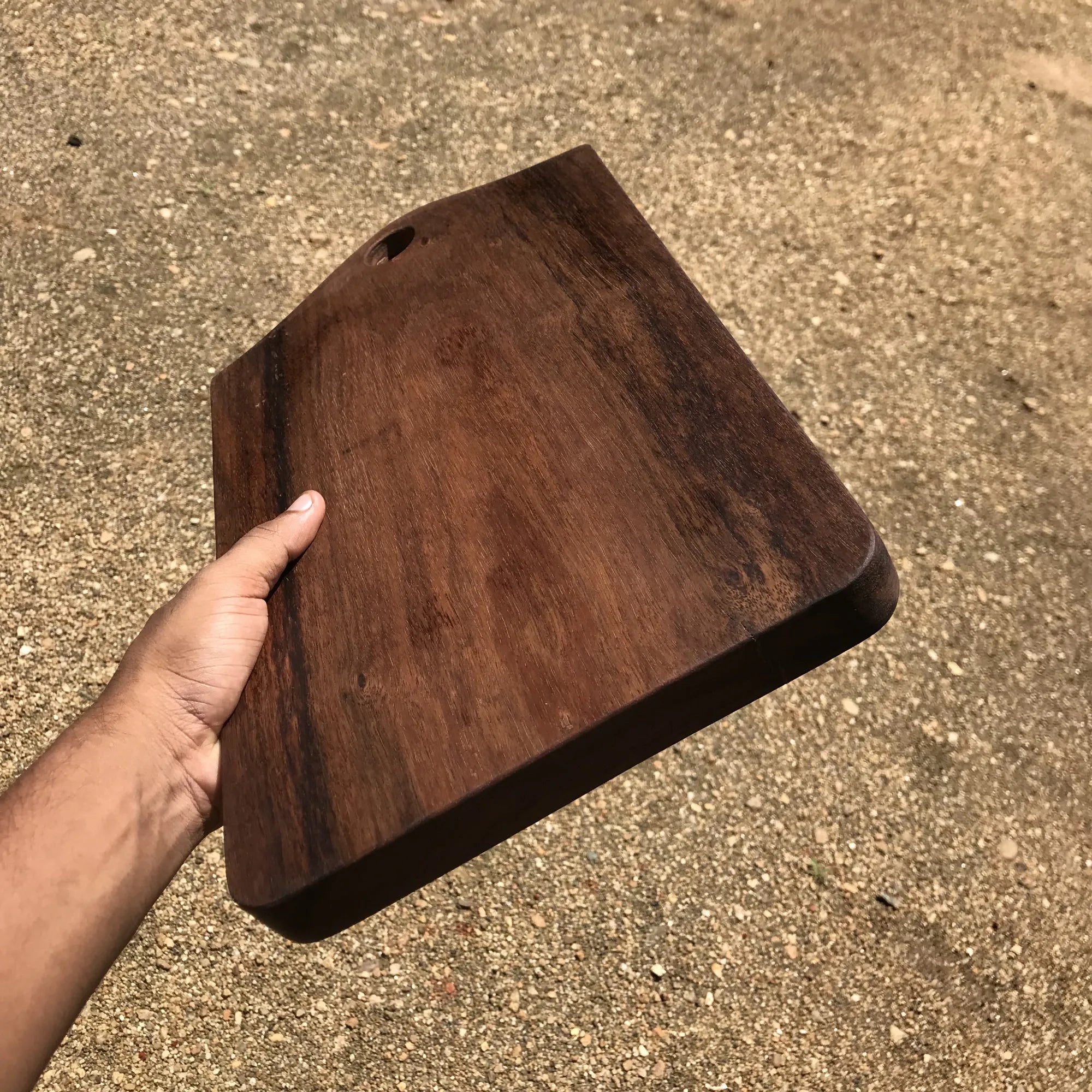 Hand holding a dark brown wood cutting board with rounded edges and a hole for hanging
